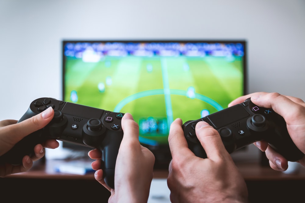 Two hands holding PlayStation controllers with a game screen in the background