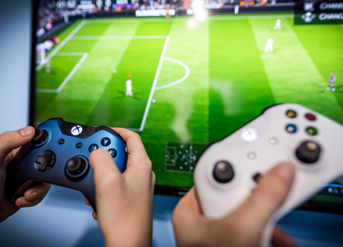 Two hands holding Xbox controllers in front of a TV showing a football game.