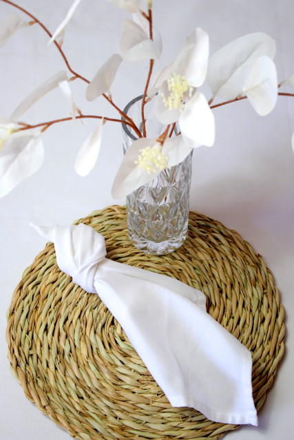 Table setting featuring white flowers in a glass vase and a white napkin on a woven placemat.