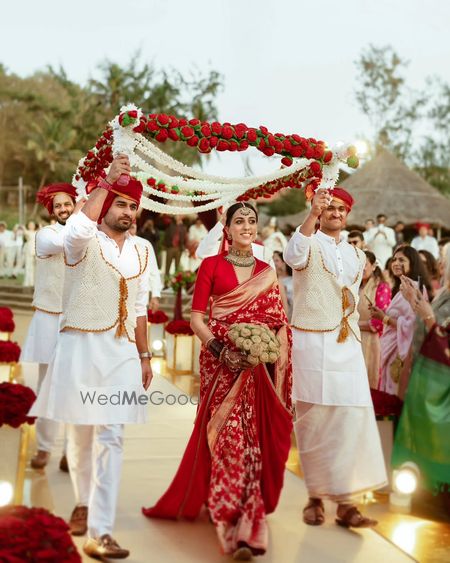 Bride in red saree with floral canopy during wedding procession.