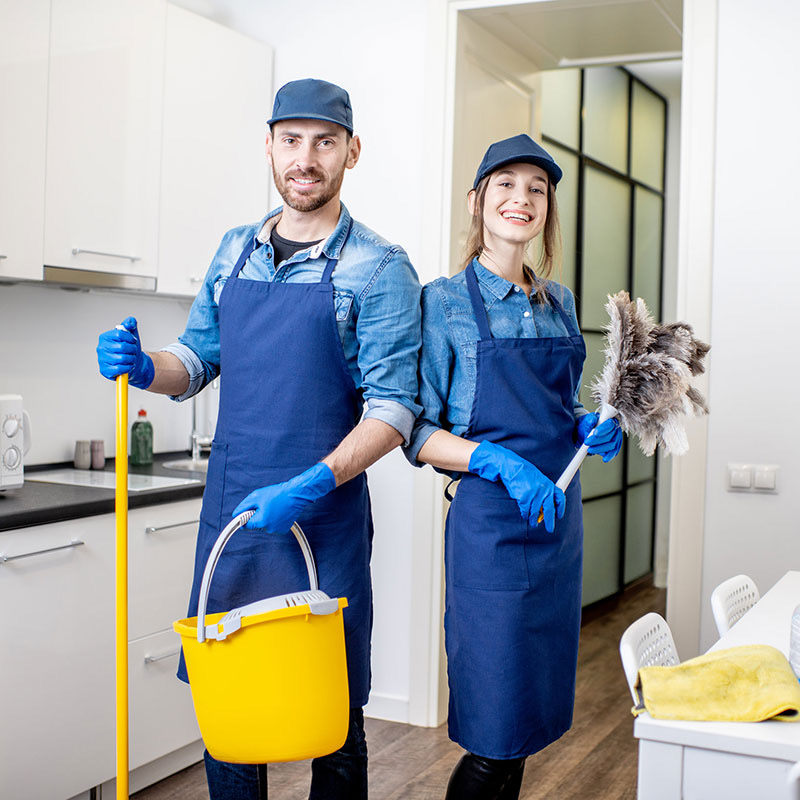 Two cleaning professionals in uniforms hold cleaning tools in a kitchen.