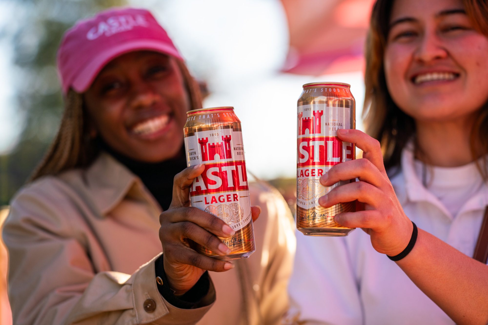 Two women toasting with Castle Lager cans outdoors.