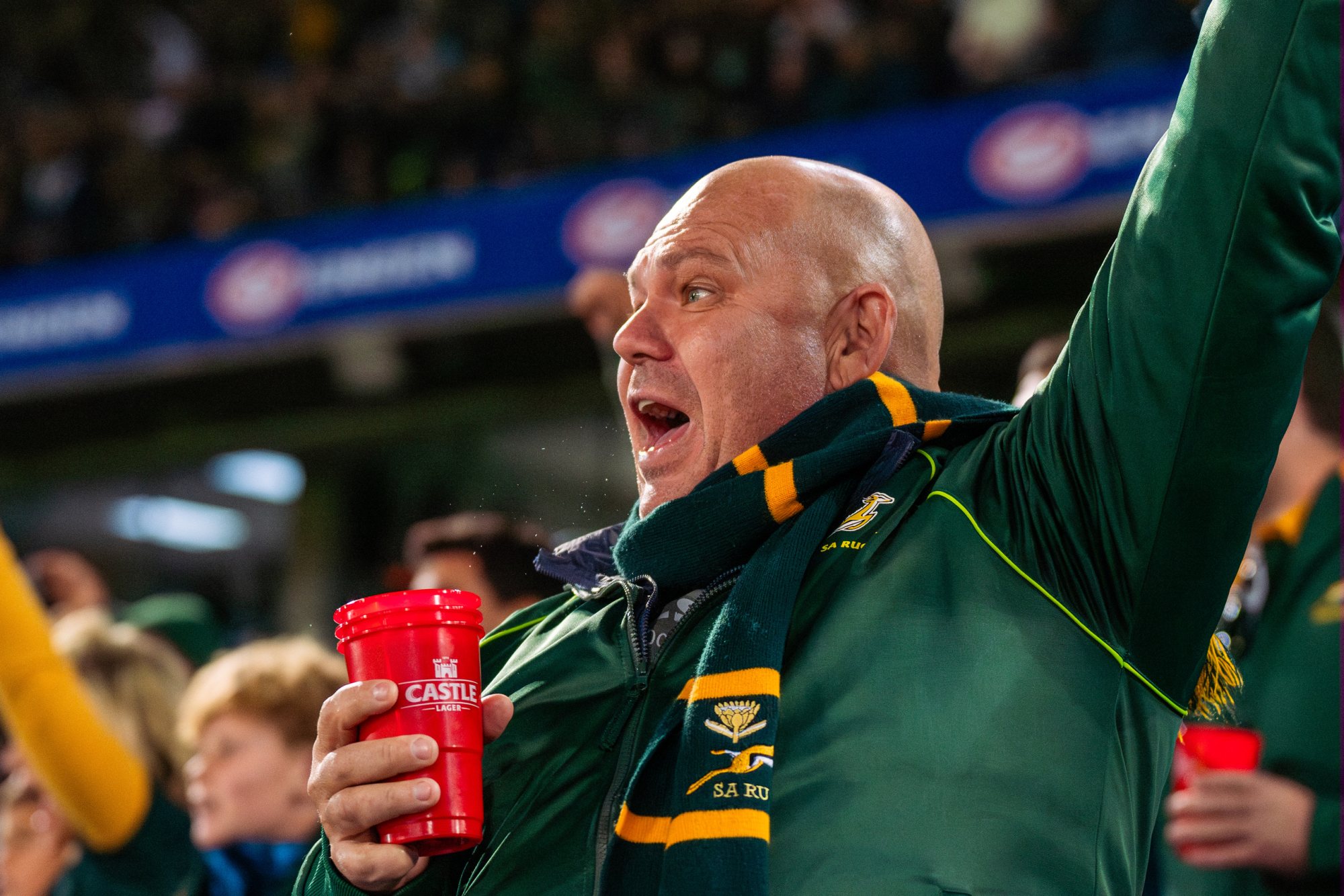 A man in a green jacket cheers with a red cup at a stadium.