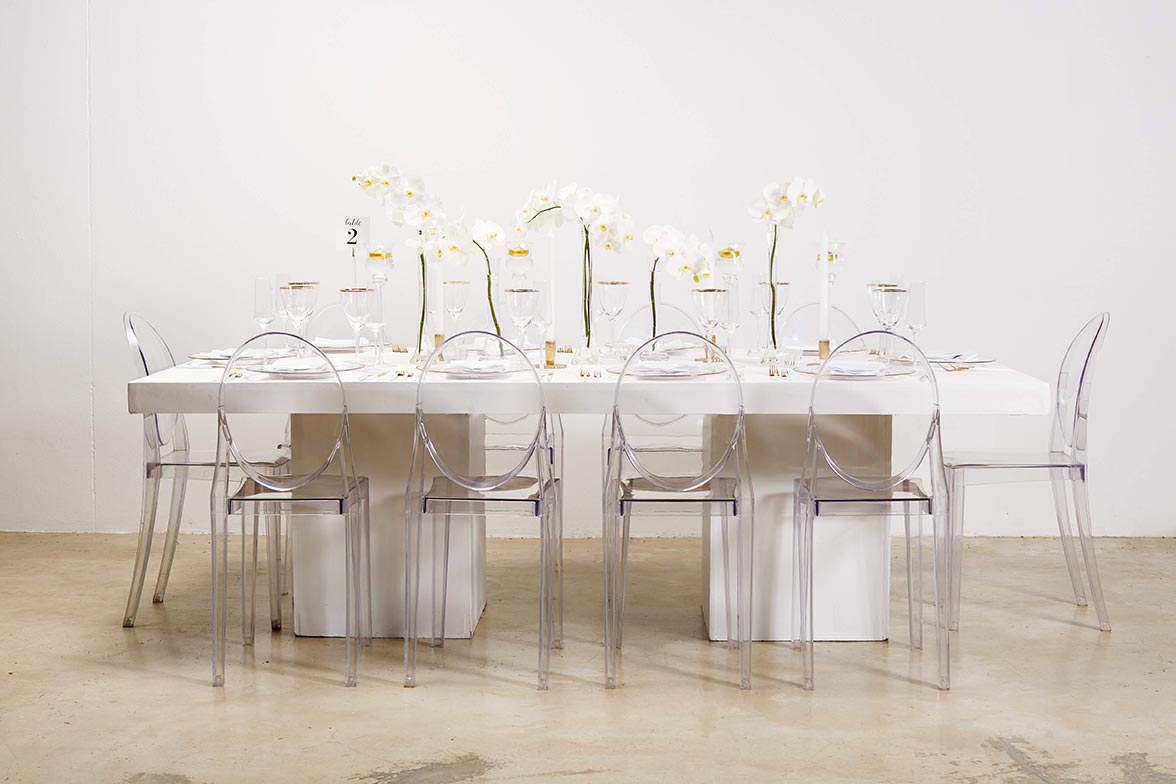 White dining table with clear chairs and orchid centerpieces.