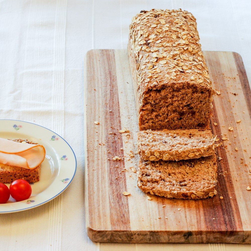 Sliced oat bread on a cutting board with toppings and tomatoes