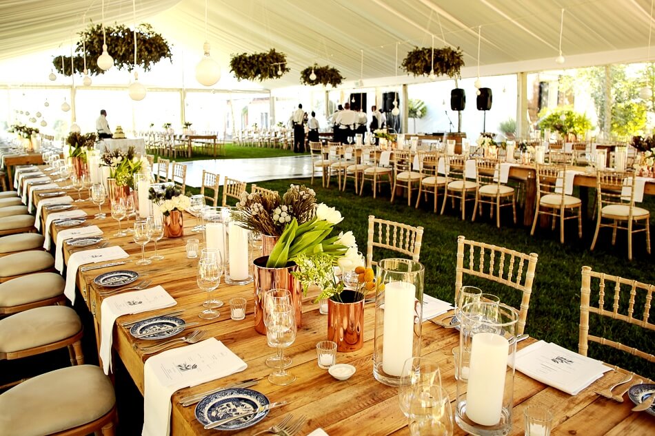 Dining setup with wooden tables, candles, and flowers in a large tent.