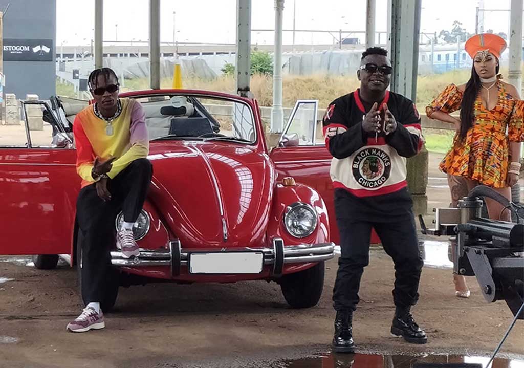 Two artists posing near a classic red Volkswagen Beetle.