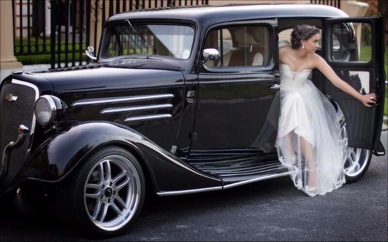 Woman in wedding dress beside a vintage black car.