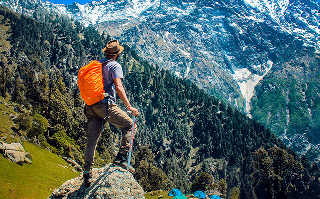 Hiker with orange backpack standing on rock, overlooking snowy mountains.