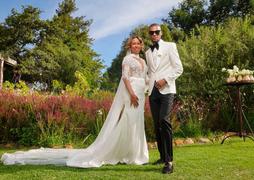 A couple posing in a garden with floral backdrop, dressed in wedding attire.