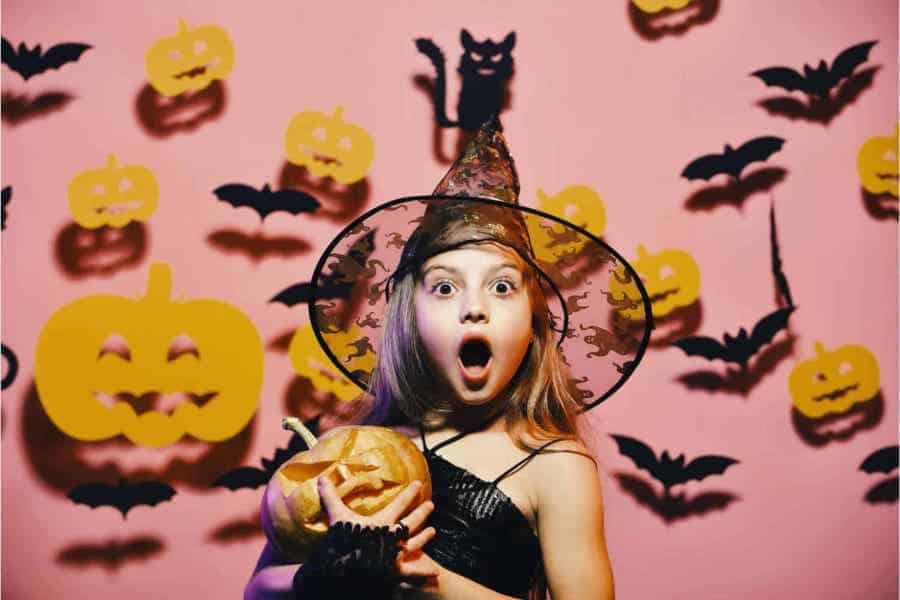 Child in witch hat holding a pumpkin with Halloween decorations in the background.
