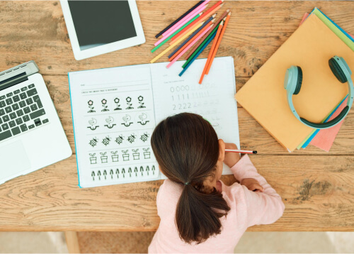 Child writing in a notebook with school supplies on a wooden table.