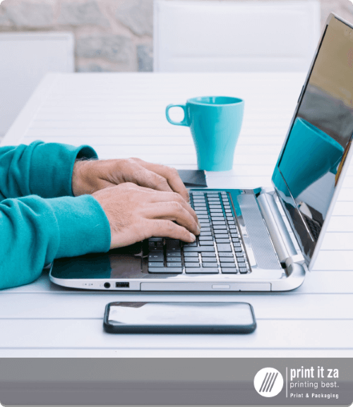 A person using a laptop with a teal mug and smartphone on a table