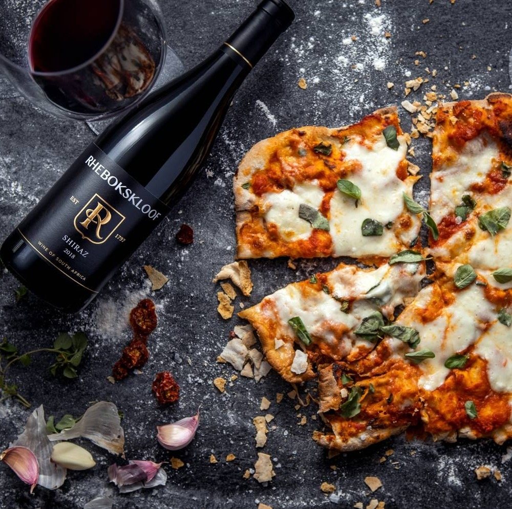 A bottle of Rhebokskloof Shiraz with a pizza on a rustic surface.