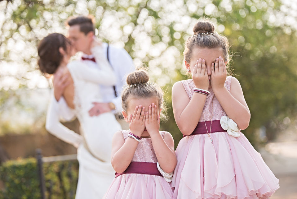 Two girls in pink dresses covering their eyes with a couple kissing in the background.