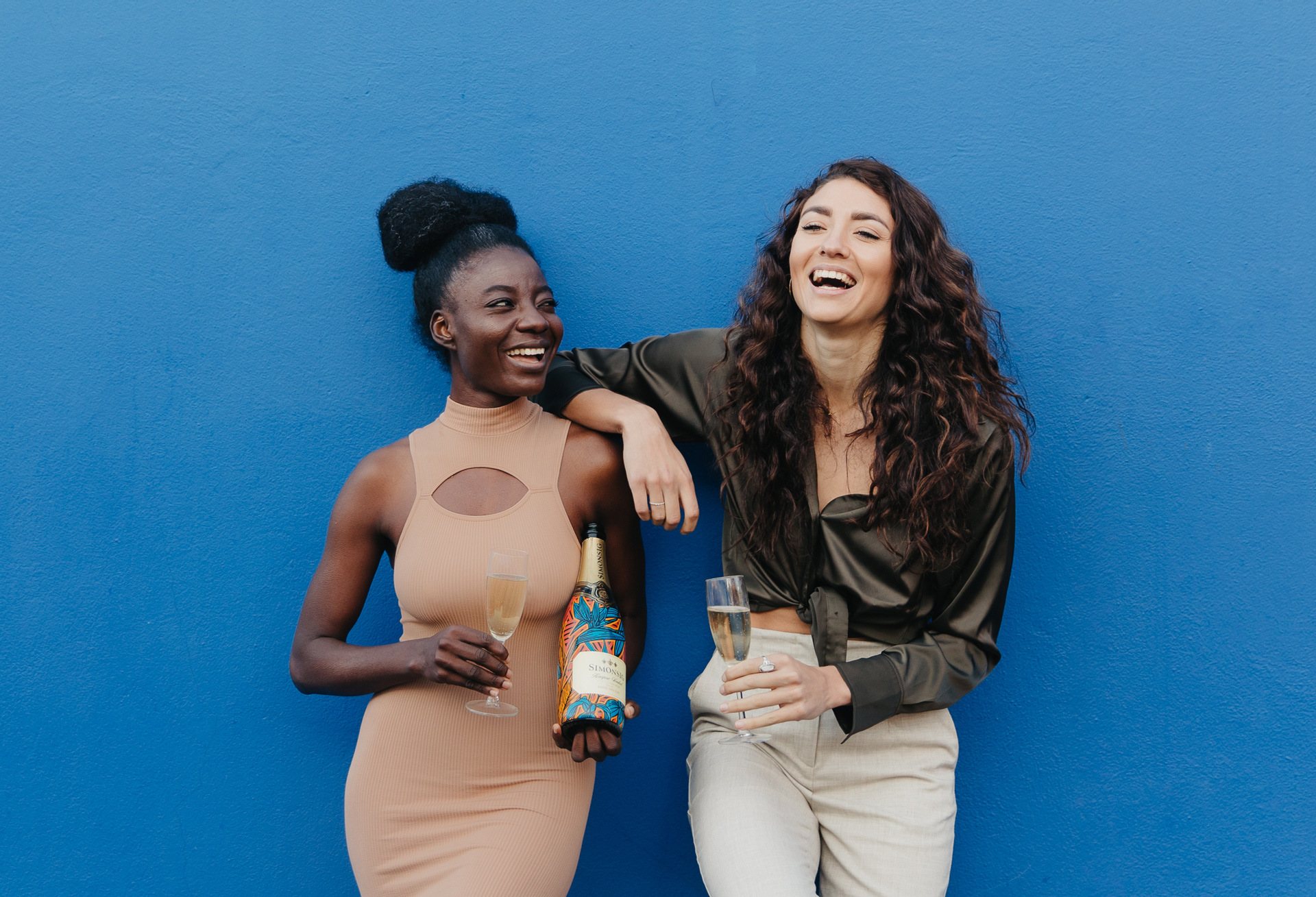 Two women holding sparkling wine, smiling against a blue wall.