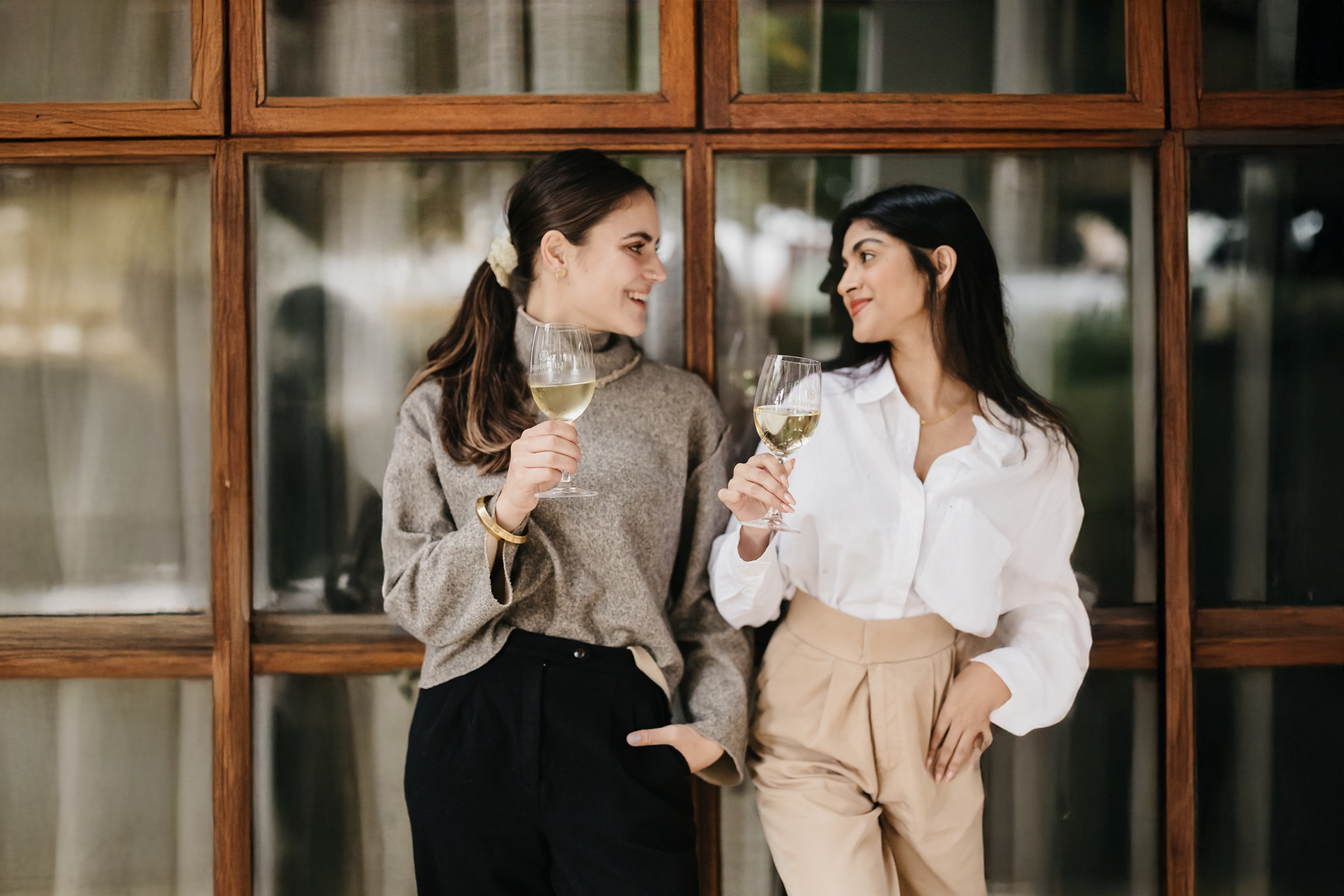 Two women holding glasses of white wine in front of wooden windows