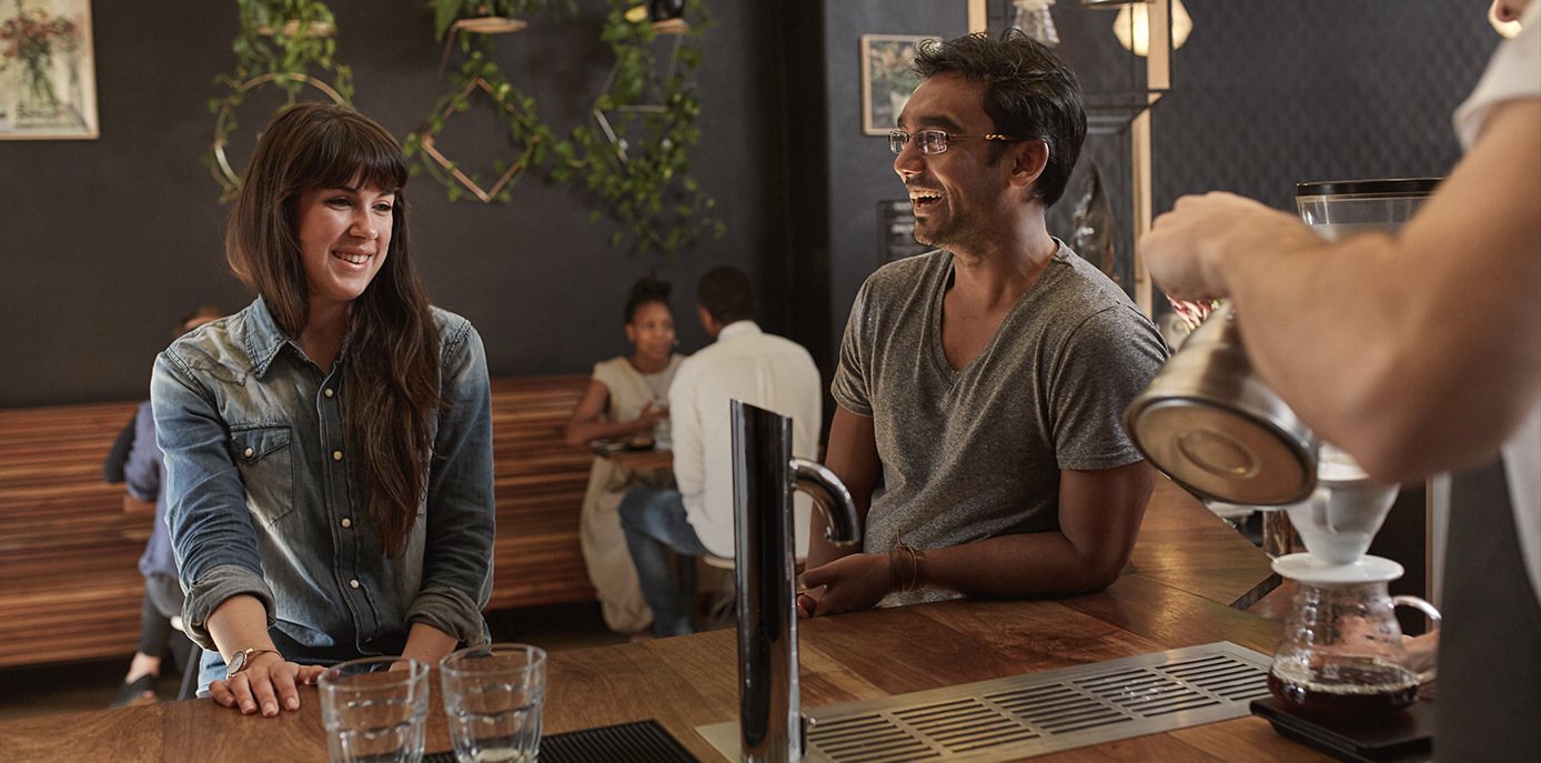 Two customers smiling at a coffee preparation demonstration in a café.