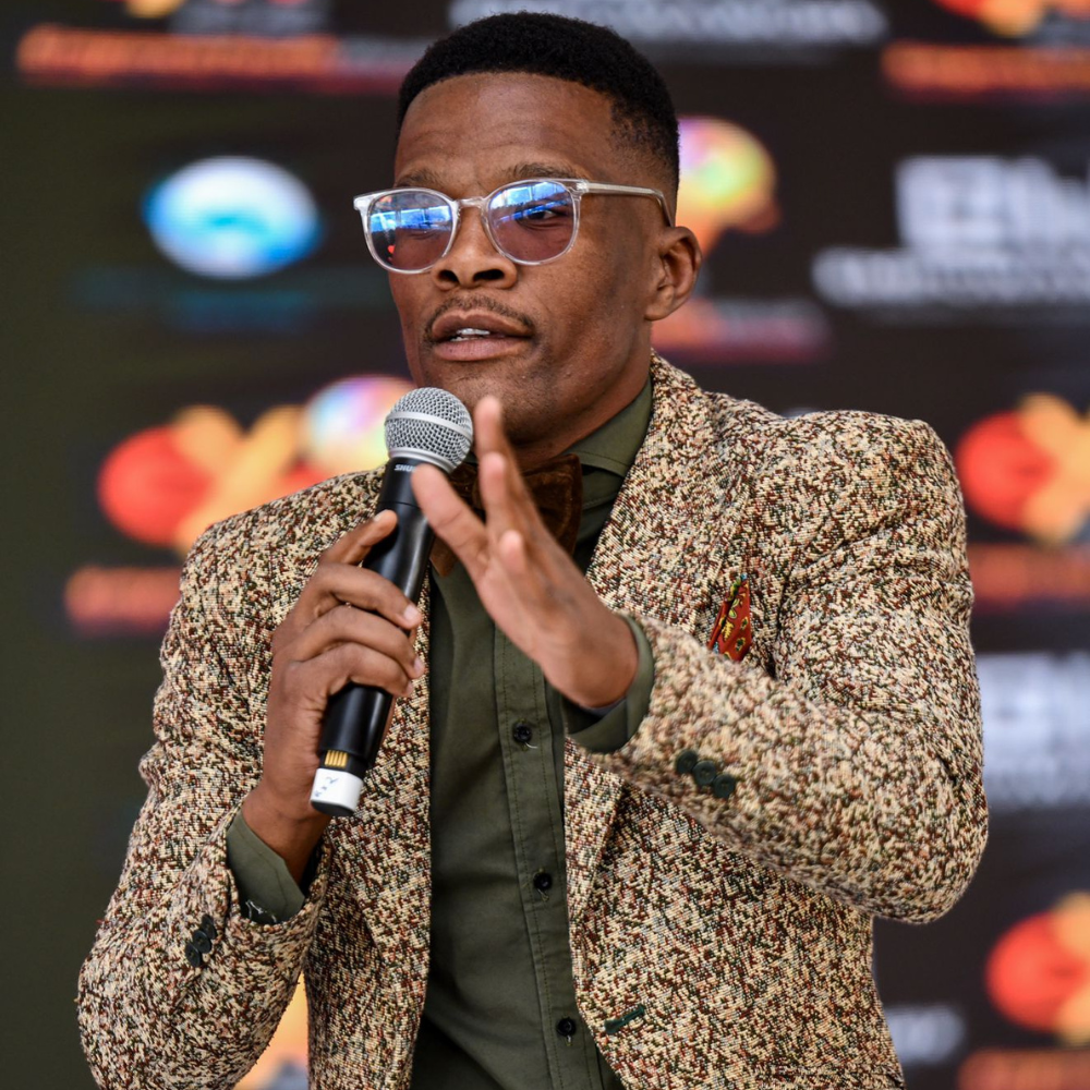 Comedian in patterned jacket speaking into a microphone on stage.