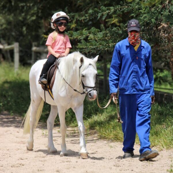 Child riding a pony led by an adult in a riding arena.