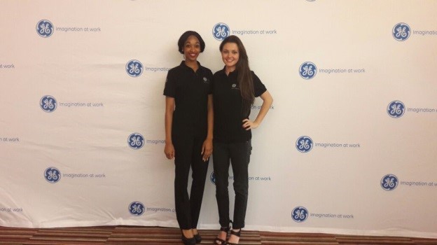 Two women in black shirts with GE logos standing by a branded backdrop.