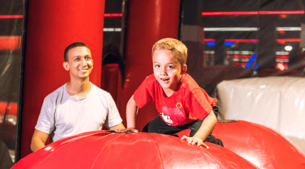 A child climbing on a red inflatable surface with an adult smiling nearby.