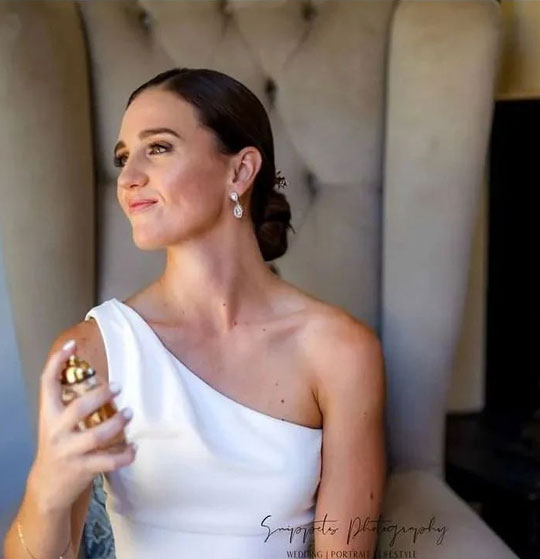 Bride in one-shoulder gown applying perfume in elegant chair