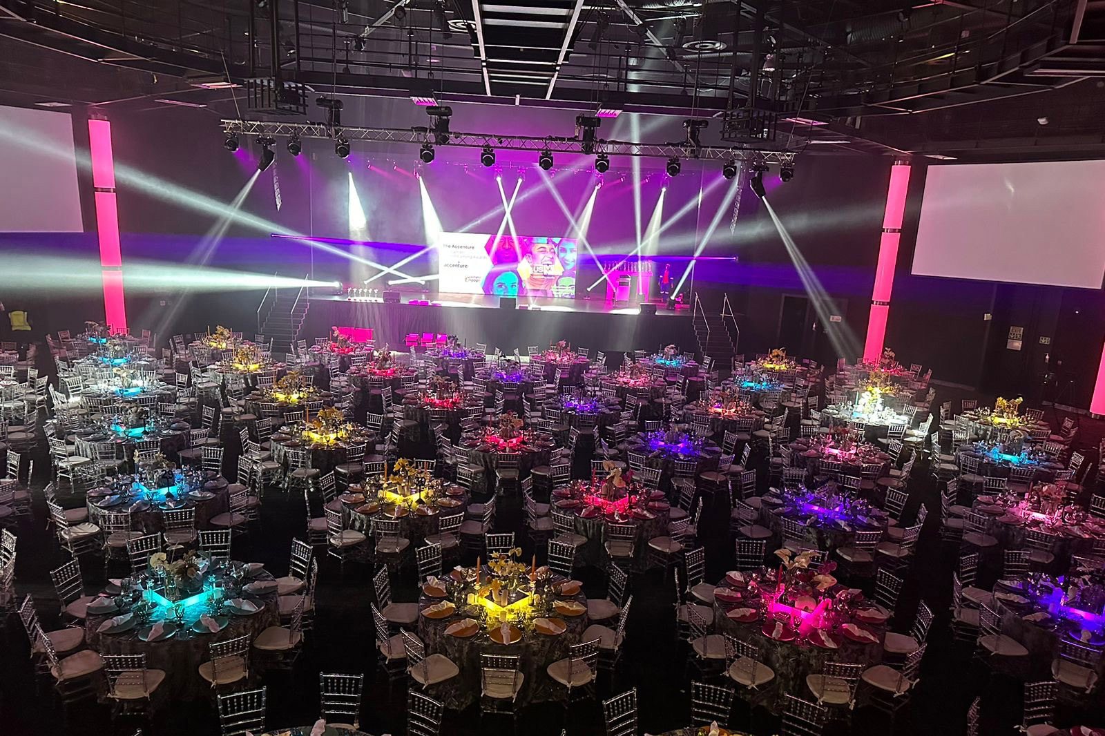 Wide view of a colorful event setup with tables and stage lighting.