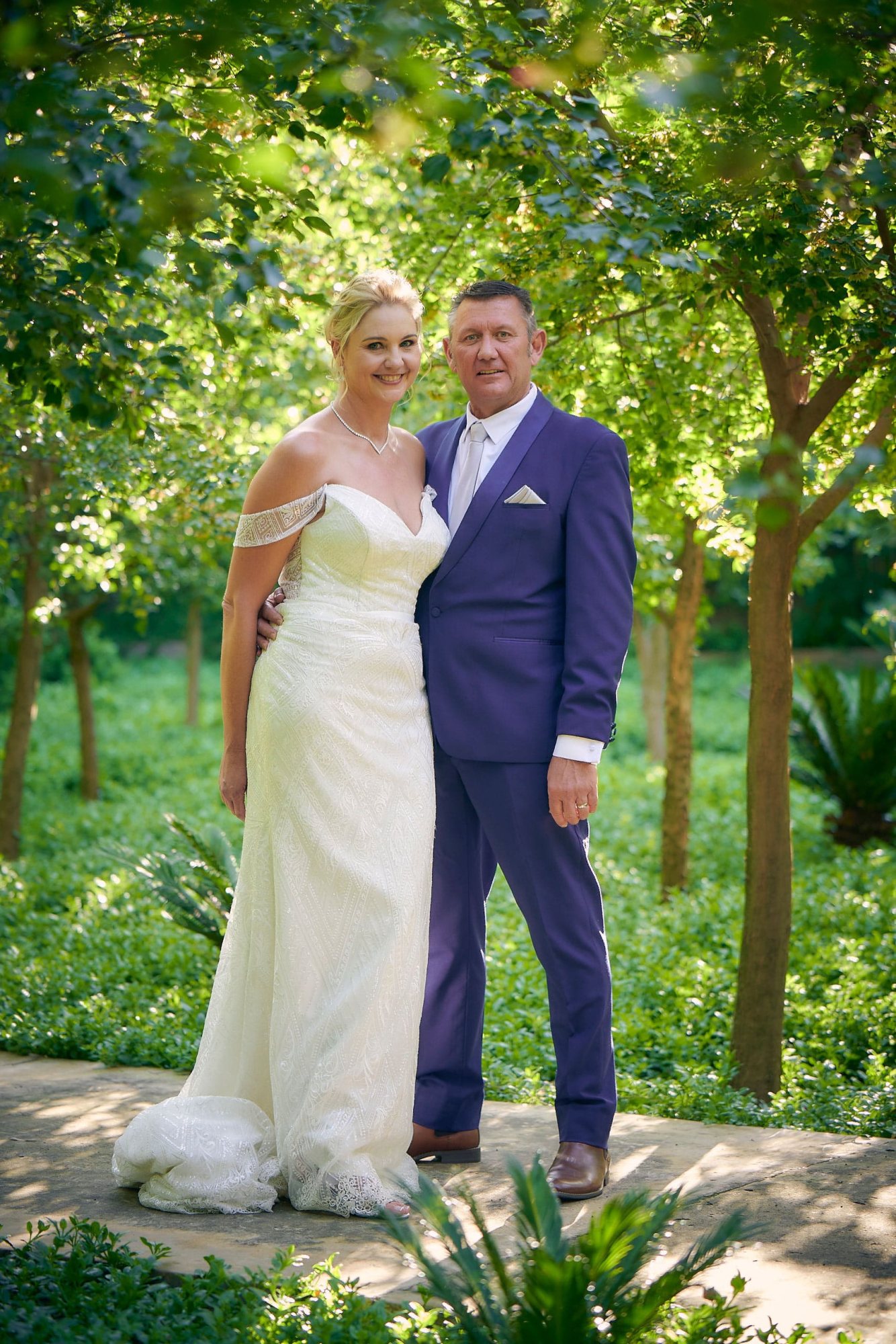 Bride in white gown and groom in purple suit in a garden.