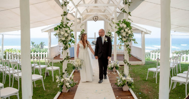 Couple walking down a wedding aisle with floral decor and ocean views.