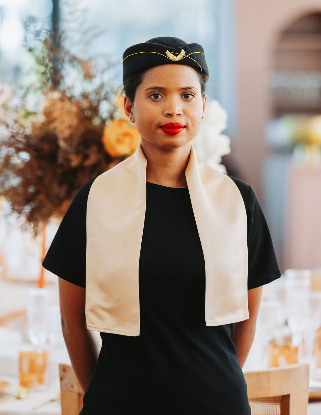 A professional hostess in a black outfit and cream scarf, poised and ready to serve.